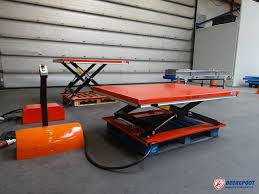 Je ziet een laagbouw rood/ oranje heftafel van Beerepoot met op de achtergrond nog een oranje heftafel en een grijs bandje in de hoek. De heftafel staat in de werkplaats van Beerepoot.